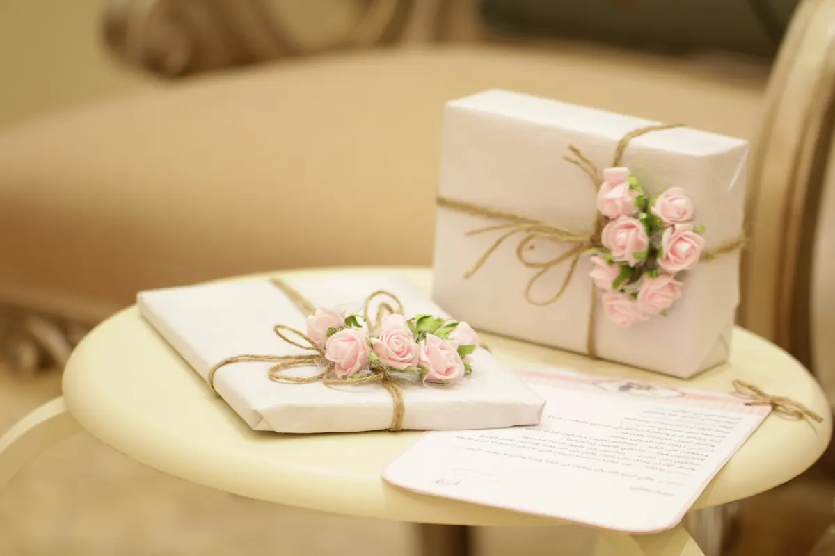 Two wrapped gifts with pink roses beside a handwritten card on a small table.